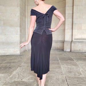 Op de afbeelding: Een vrouw draagt een zwart, off-shoulder top met een glinsterende textuur en een zwarte, asymmetrische rok. De rok heeft een geplooide detail aan de achterkant.
