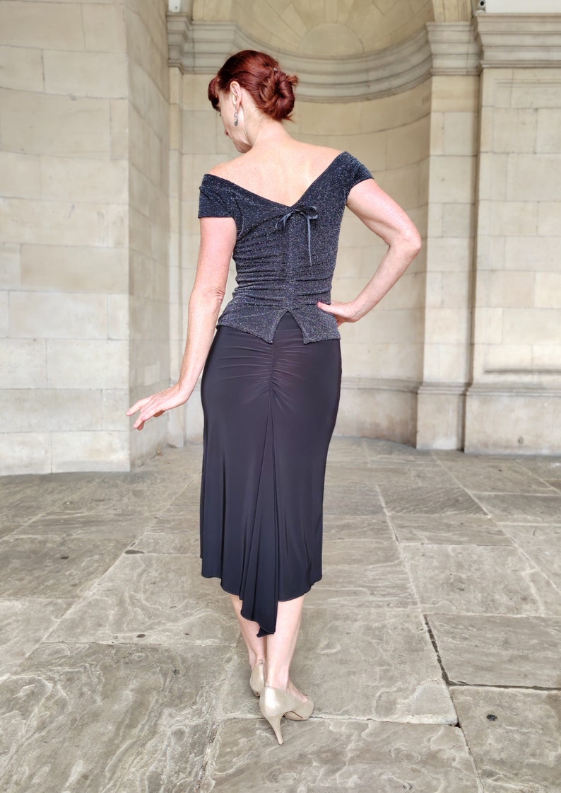 Op de afbeelding: Een vrouw draagt een zwart, off-shoulder top met een glinsterende textuur en een zwarte, asymmetrische rok. De rok heeft een geplooide detail aan de achterkant.