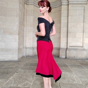 Op de afbeelding: Een vrouw draagt een zwart off-shoulder top met een glinsterende textuur en een rode rok met een zwarte bies. De rok is strak om de taille en loopt uit naar beneden.
