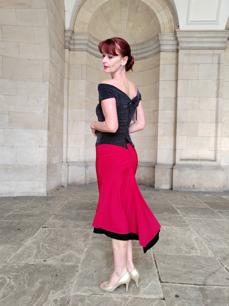 Op de afbeelding: Een vrouw draagt een zwart off-shoulder top met een glinsterende textuur en een rode rok met een zwarte bies. De rok is strak om de taille en loopt uit naar beneden.