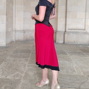 Op de afbeelding: Een vrouw draagt een zwart off-shoulder top met een rode rok. De rok heeft een zwarte bies en is zwierig. Ze draagt gouden hakken.