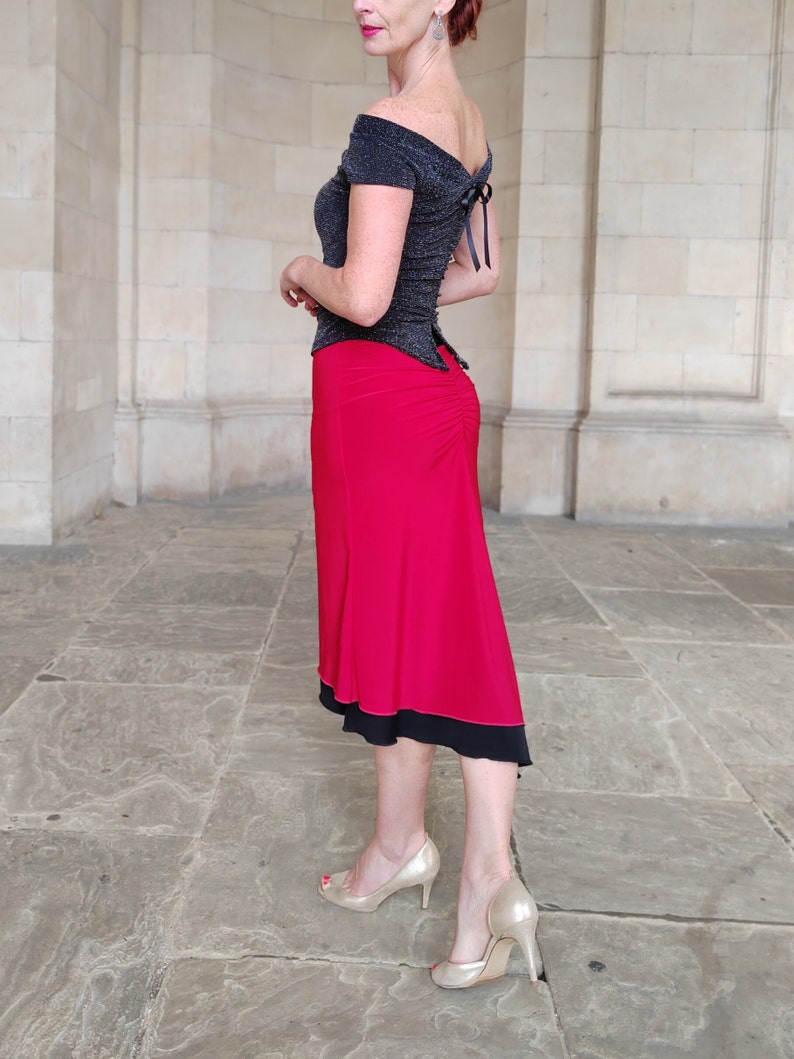 Op de afbeelding: Een vrouw draagt een zwart off-shoulder top met een rode rok. De rok heeft een zwarte bies en is zwierig. Ze draagt gouden hakken.