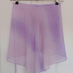 Puede incluir: Una falda envolvente lavanda con un delicado estampado floral, colgada de una percha. La falda tiene un dobladillo asimétrico y lazos largos. La tela pasa de un lavanda más oscuro en la parte superior a un tono más claro en la inferior.