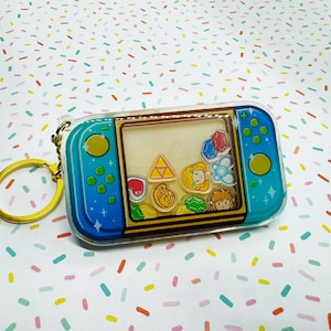 May include: A blue and teal keychain resembling a game console, with a gold key ring. The keychain features a clear window with colorful game-themed charms inside. The background is a white surface with colorful sprinkles.