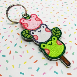 May include: A novelty keychain with three embroidered frog-shaped treats. The frogs are pink, white, and green, each with a smiling face and leaf details. The keychain has a silver metal ring.
