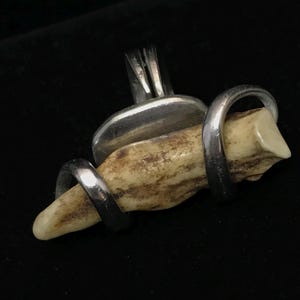 May include: A ring featuring a horizontal, textured, off-white and brown bone-like fragment secured by two polished silver-toned metal loops. The metal band is attached to a flat, rectangular silver-toned plate behind the central fragment.