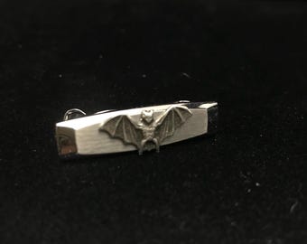 May include: A silver tie bar with a bat design. The bat is in a flying position with its wings spread out.