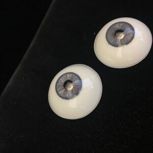 May include: Two realistic-looking glass eyes with a gray iris and a silver fleck in the center. The eyes are set against a black background.