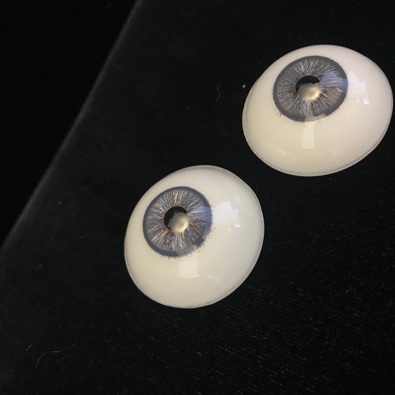 May include: Two realistic-looking glass eyes with a gray iris and a silver fleck in the center. The eyes are set against a black background.
