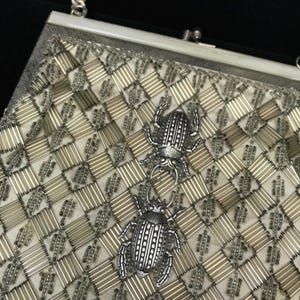 Op de afbeelding: Een vintage zilverkleurige handtas met een geometrisch patroon van zilveren staven en een crèmekleurige stoffen achtergrond. Twee decoratieve zilveren keverversieringen zijn bevestigd aan de voorkant van de tas. De tas heeft een zilveren sluiting en ketting.