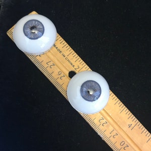 May include: Two white plastic doll eyes with a blue iris and a silver pupil. The eyes are about 1 inch in diameter and are sitting on a wooden ruler.