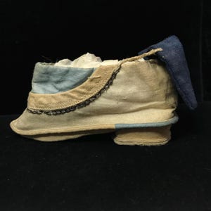 May include: A vintage-style shoe in cream, blue, and tan. The shoe features a small heel, a blue accent on the toe, and a blue fabric flap at the back. The shoe is trimmed with a decorative border.