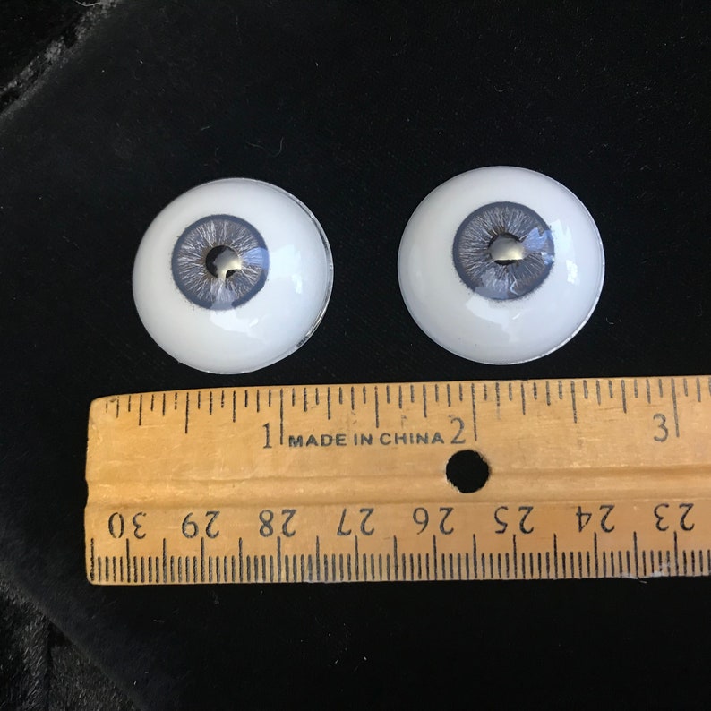 May include: Two white doll eyes with a gray iris and a silver pupil. The eyes are approximately 1 inch in diameter. A ruler is in the background for size reference.
