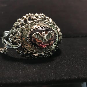 Op de afbeelding: Een zilveren armband met een sierlijk ontwerp heeft een grote, gefacetteerde, paarse edelsteen met een zilveren vleermuisontwerp in het midden.