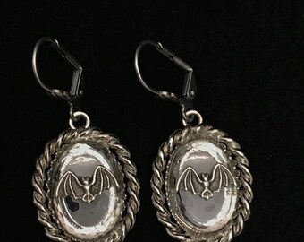 May include: A pair of silver-toned earrings with a bat design. Each earring features an oval-shaped pendant with a bat in the center, surrounded by a twisted rope-like border. The earrings have a lever-back closure.