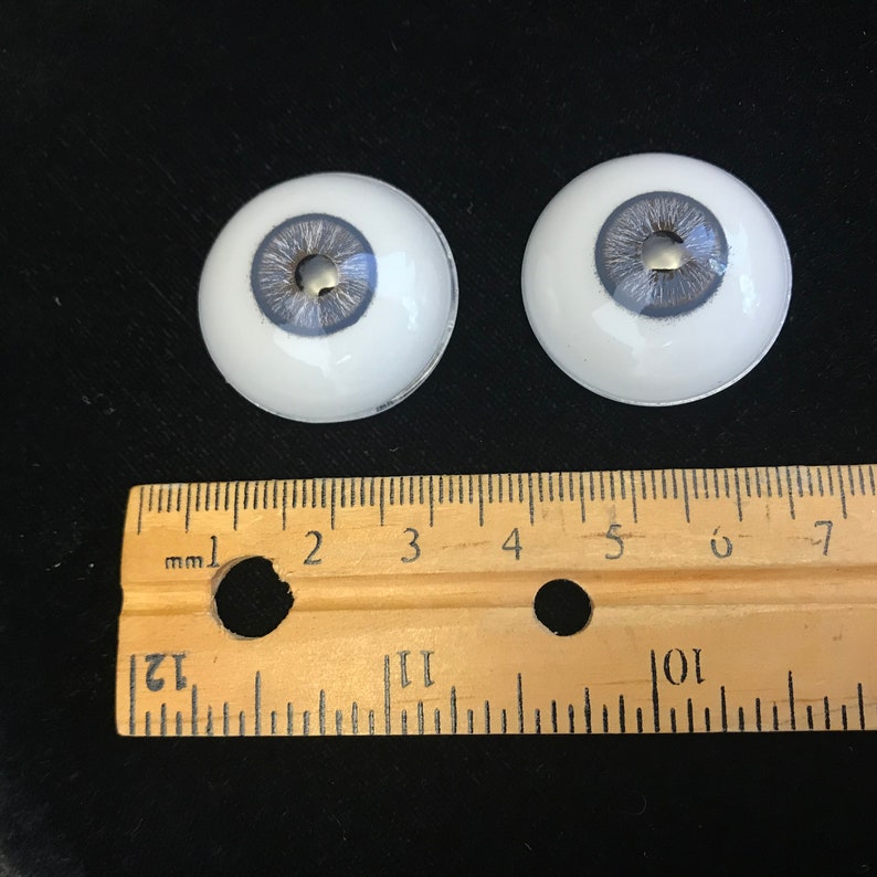 May include: Two white plastic doll eyes with a gray iris and a silver pupil. The eyes are approximately 1 inch in diameter.