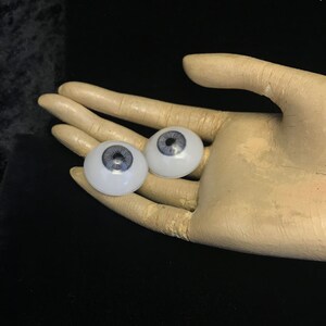 May include: Two realistic-looking glass eyes with gray irises and white scleras. The eyes are sitting on a pale beige hand.