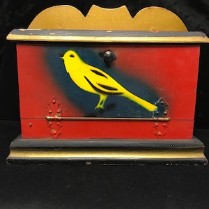Op de afbeelding: Een vintage houten kist met een rode voorkant met een gele vogelontwerp. De kist heeft een goudkleurige boven- en onderkant, met zwarte rand. Decoratieve scharnieren en een kleine knop zijn zichtbaar aan de voorkant.