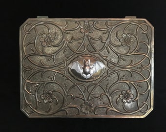May include: A rectangular, silver-toned box with an ornate floral and vine pattern. A central oval features a detailed bat design. The box has a hinged lid and is likely made of metal. The overall aesthetic is gothic and decorative.