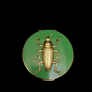 May include: A round, green compact with a gold-colored beetle design in the center. The beetle is highly detailed with textured wings and legs. The compact has a gold-colored rim and a black background.