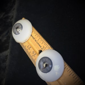 May include: Two white plastic doll eyes with gray irises and black pupils. The eyes are placed on a wooden ruler marked with inches and the text "Made in China".