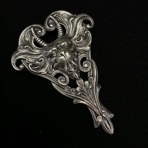 May include: A silver-toned, ornate, decorative brooch with a detailed, stylized, animal head in the center.