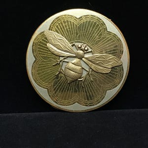 May include: A round, gold-toned compact with a detailed bee design. The bee is centered on a gold floral pattern, surrounded by a silver-colored border. The compact is likely made of metal and has a vintage or antique aesthetic.