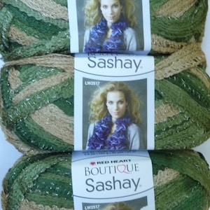 May include: Three skeins of Red Heart Boutique Sashay yarn in shades of green, tan, and silver. Each skein has a label with a woman's portrait and the text "Sashay". The yarn is ideal for knitting and crochet projects.