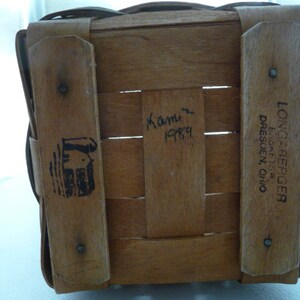 May include: A woven wooden basket with a rectangular shape. The basket has wooden supports and a handle. The text "LONGABERGER, DRESDEN, OHIO" is printed on one of the wooden supports. The basket also has a black logo and the handwritten text "Kami 1989".