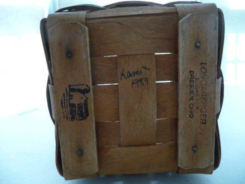 May include: A woven wooden basket with a rectangular shape. The basket has wooden supports and a handle. The text "LONGABERGER, DRESDEN, OHIO" is printed on one of the wooden supports. The basket also has a black logo and the handwritten text "Kami 1989".