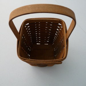 May include: A square, woven basket with a wooden handle. The basket is made of light brown woven strips, with a matching handle arching over the top. The basket's interior is visible, showing the woven pattern and the empty space within.