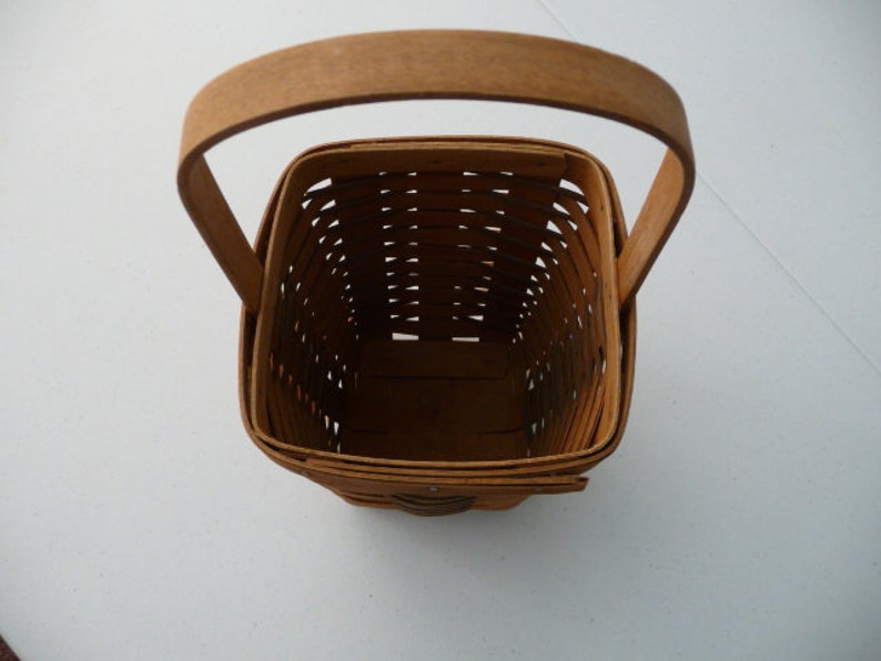 May include: A square, woven basket with a wooden handle. The basket is made of light brown woven strips, with a matching handle arching over the top. The basket's interior is visible, showing the woven pattern and the empty space within.
