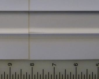 May include: A clear, rectangular glass rod, approximately 13 inches long, lies on a white surface next to a ruler. The rod has a smooth, polished appearance and is transparent, allowing light to pass through. The object is likely used for scientific or decorative purposes.