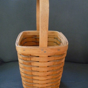 May include: A light brown woven basket with a rectangular base and a curved handle. The basket is made of thin, interwoven strips of wood, creating a textured surface. The handle is attached to the top of the basket with small metal rivets.