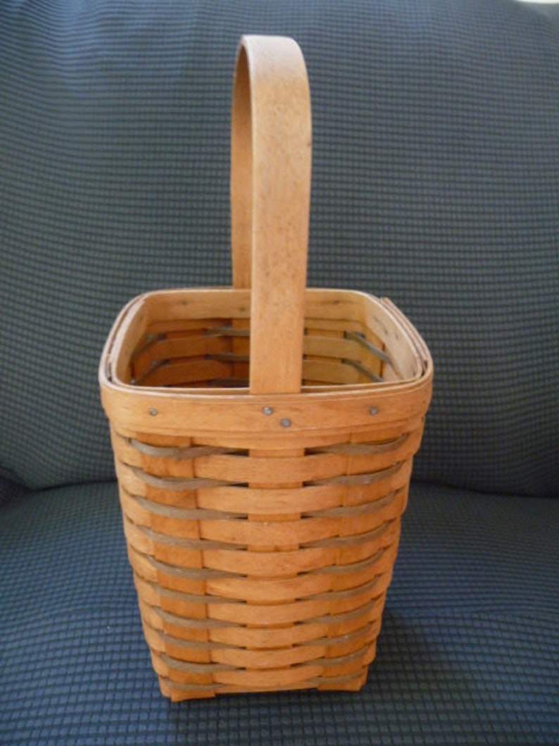May include: A light brown woven basket with a rectangular base and a curved handle. The basket is made of thin, interwoven strips of wood, creating a textured surface. The handle is attached to the top of the basket with small metal rivets.