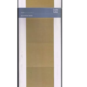 May include: A packaged accordion book with a textured, tan-colored interior. The packaging includes text that reads "QuickKutz dies can both cut and emboss" and "Cookie Cutter." The book is designed for crafting and scrapbooking.