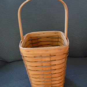 May include: A light brown, woven basket with a curved handle. The basket is rectangular with a flared shape and is made of thin, horizontal strips of wood. The handle is also made of wood and arches over the top of the basket.