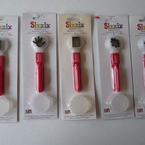 May include: Five Sizzix PaddlePunch hand punches in their original packaging. Each punch has a red handle, a white head with a different shape, and a white base. The packaging includes the Sizzix logo and product information.