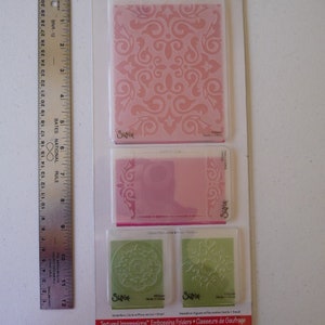 May include: A package of Sizzix Textured Impressions embossing folders. The package includes three different designs: a large pink floral pattern, a pink border design, and two green medallion designs. A metal ruler is on the left side of the image.