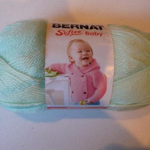 May include: A skein of Bernat Softee Baby yarn in a soft, pale green color. The label features a smiling baby wearing a pink jacket. The yarn is tightly wound and appears soft, ideal for baby projects.