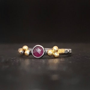 May include: A silver ring with a red gemstone and three gold beads on each side.