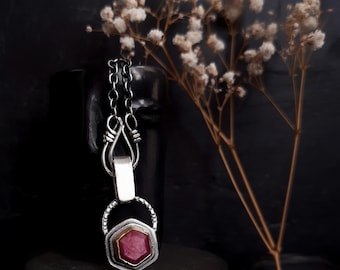 May include: A silver necklace with a hexagonal pendant featuring a red gemstone set in gold. The pendant is surrounded by a silver frame with a textured design.