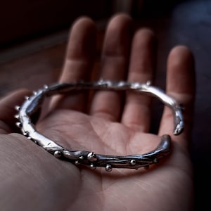 May include: A silver-colored, open-ended bracelet with a textured, organic design. The bracelet features small, rounded protrusions and a rough, uneven surface. The jewelry is held in an open hand, showcasing its unique, handcrafted appearance.