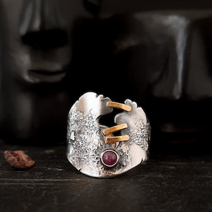 May include: A silver ring with a textured surface and gold accents. The ring has a pink gemstone set in the centre.