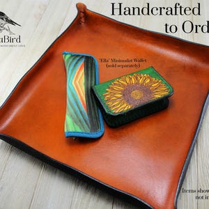 May include: A handcrafted leather tray in a warm brown hue, with a black trim. The tray holds a colorful wallet and a green wallet with a sunflower design. The text "Handcrafted to Order" is visible.