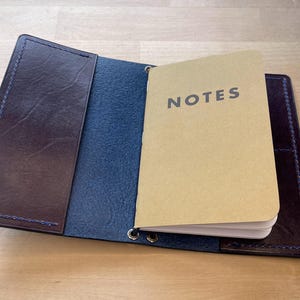 May include: A dark purple leather notebook cover with blue stitching, featuring a textured blue interior lining and metal eyelets. A tan paper notebook with the word NOTES printed in black on the front cover is inserted into the leather sleeve.