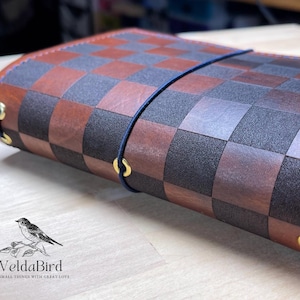May include: A leather journal cover featuring a brown and dark charcoal checkerboard pattern. The cover is secured with a dark blue elastic cord threaded through gold-toned metal eyelets. The logo VeldaBird with a bird illustration is visible.