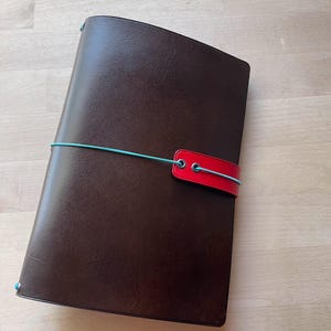 May include: A dark brown rectangular notebook cover with a smooth, matte finish. A bright turquoise elastic cord wraps horizontally around the cover, securing a rectangular red leather-like closure tab with two metal eyelets at the center.