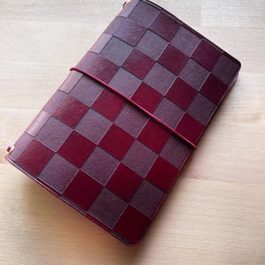 May include: A rectangular journal cover featuring a checkered pattern of alternating dark burgundy and deep maroon squares. The cover is secured by a thin, dark red elastic band that wraps horizontally around the center of the notebook.