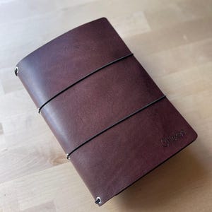 Pode incluir: Uma capa de diário em couro castanho escuro com um acabamento mate suave e duas faixas elásticas pretas finas enroladas horizontalmente à volta do exterior. A palavra Chickee está gravada no canto inferior direito e são visíveis ilhós de metal prateado.